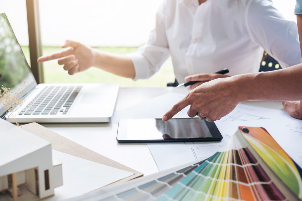 A close-up of design professionals collaborating at a conference table, pointing at a laptop, a digital tablet, architectural blueprints, and a color swatch wheel.