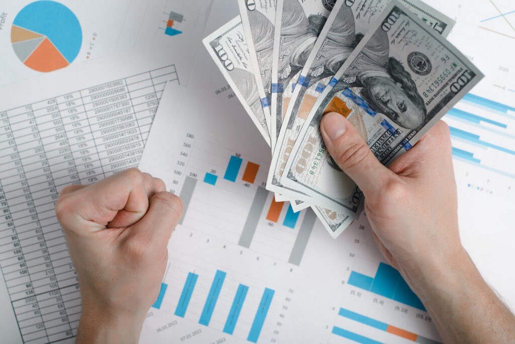 A top-down view of a business professional holding a fan of one-hundred-dollar bills in one hand and making a victorious fist with the other, positioned over financial performance charts, bar graphs, and spreadsheets analyzing business revenue data.
