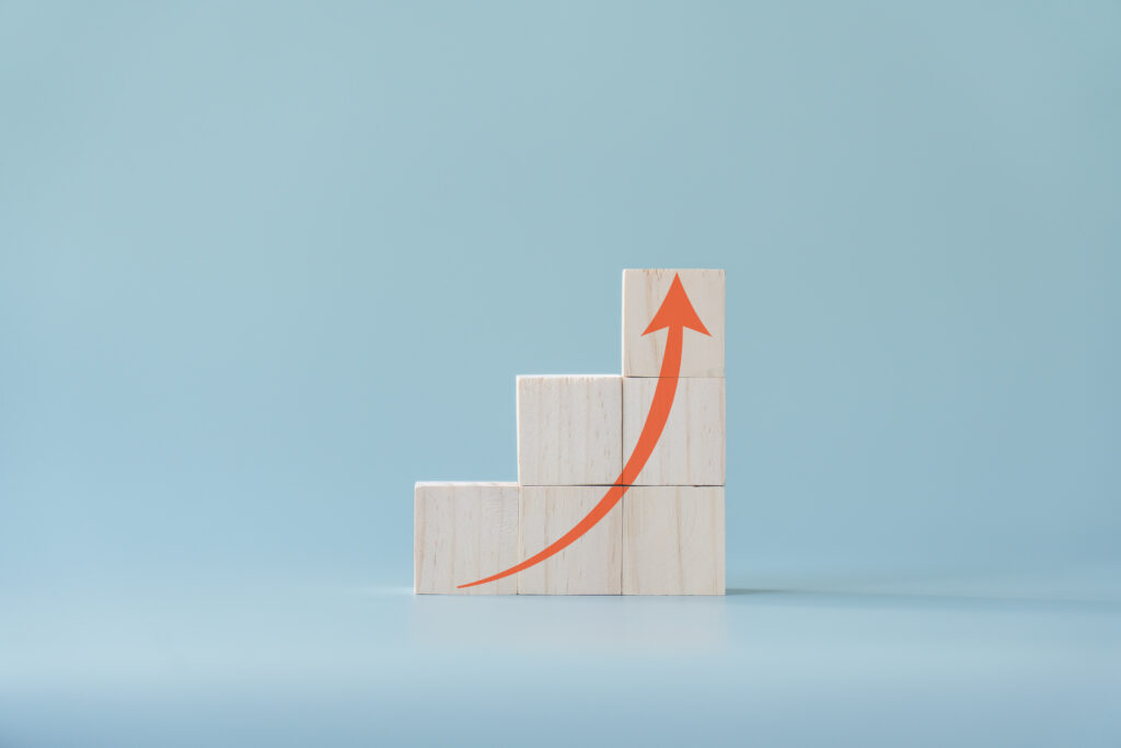 Six plain wooden toy blocks arranged in a three-step ascending staircase pattern against a solid light blue background, featuring a smooth red arrow painted across the front that curves upward to point at the highest block.