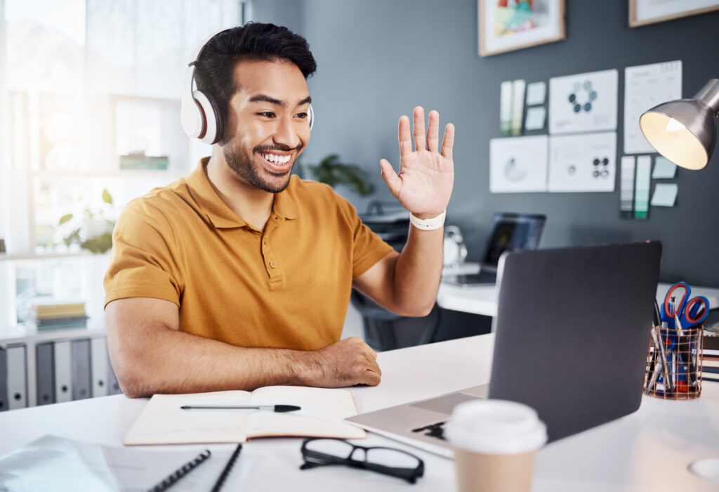 A smiling young male business professional wearing large white headphones sits at a tidy office desk and waves at his laptop screen during a video call, with an open notebook, glasses, and a coffee cup on the desk in front of him.