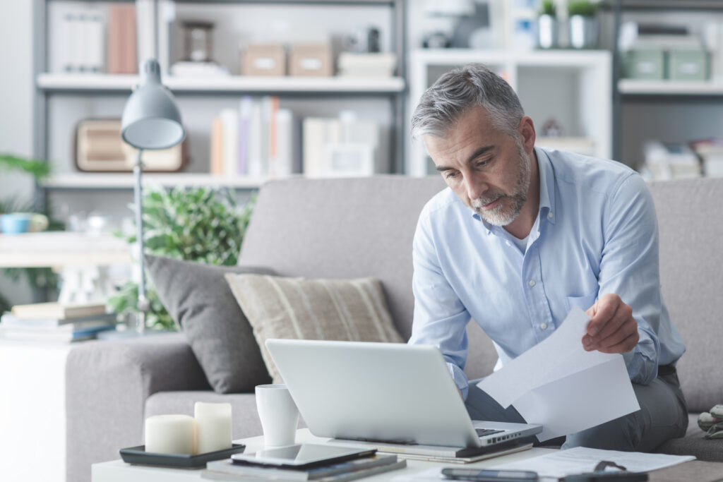 A remote healthcare billing professional reviews physical documents while working on a laptop from a home office setting.