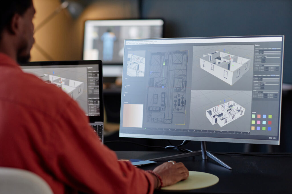A male architect from a back-shoulder view working on a 3D building layout and interior architectural model using Revit or CAD software on multiple computer monitors.