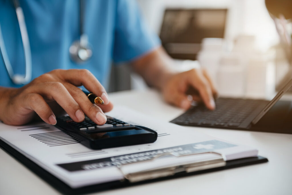 Close-up of a healthcare professional in blue scrubs using a calculator and keyboard to audit hospital revenue cycle data.