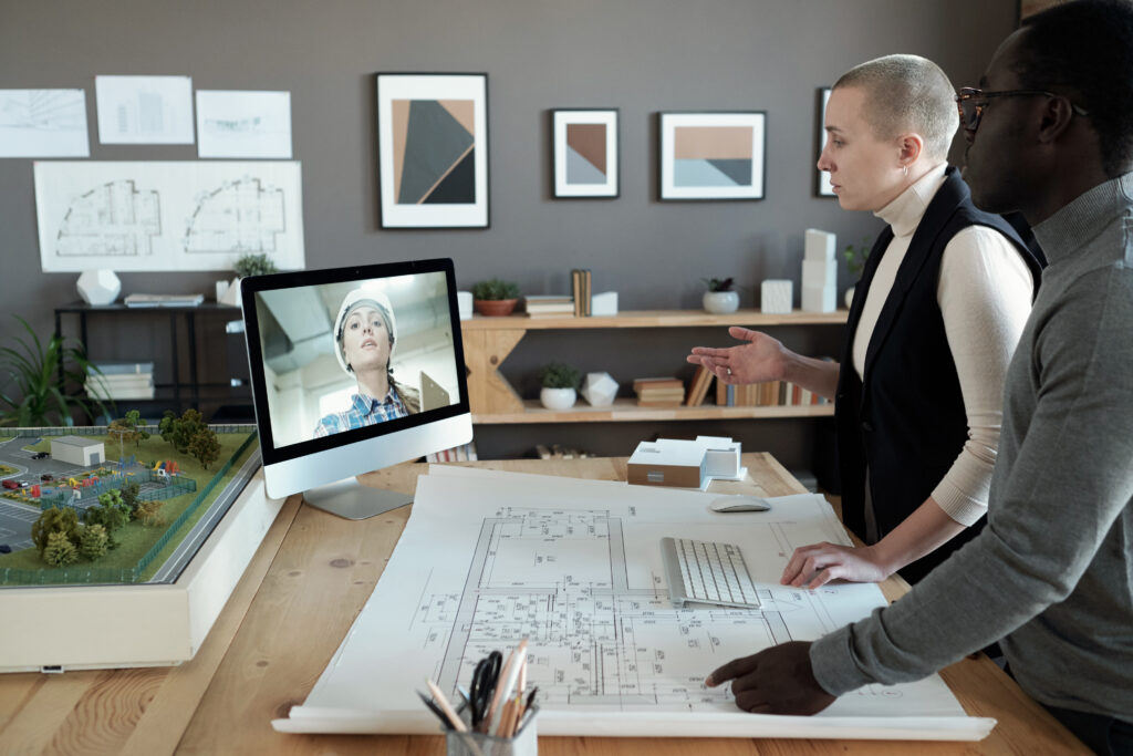 Two architects in an office reviewing printed building plans on a large table while collaborating on a live video call with a remote team member on a computer screen.