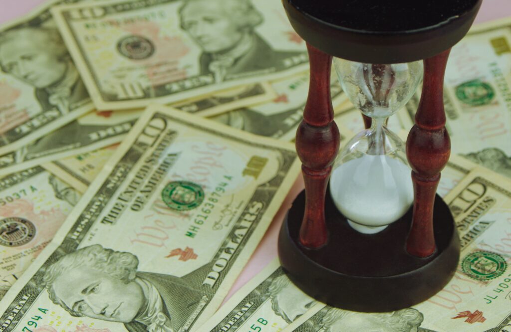 A close-up, high-angle view of a wooden hourglass with white sand running through it, sitting on top of a scattered pile of United States ten-dollar bills, representing the concept of time and money in business.