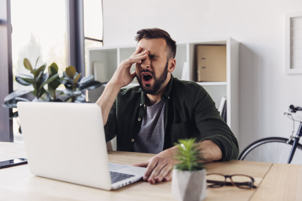 An exhausted male business professional with a beard sits at a wooden desk in a bright office, rubbing his face and yawning widely while looking at an open laptop screen.