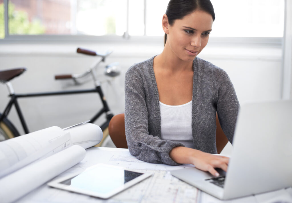 A female designer smiling and working on her laptop in a bright, modern studio with a digital tablet and rolled architectural blueprints on her desk.