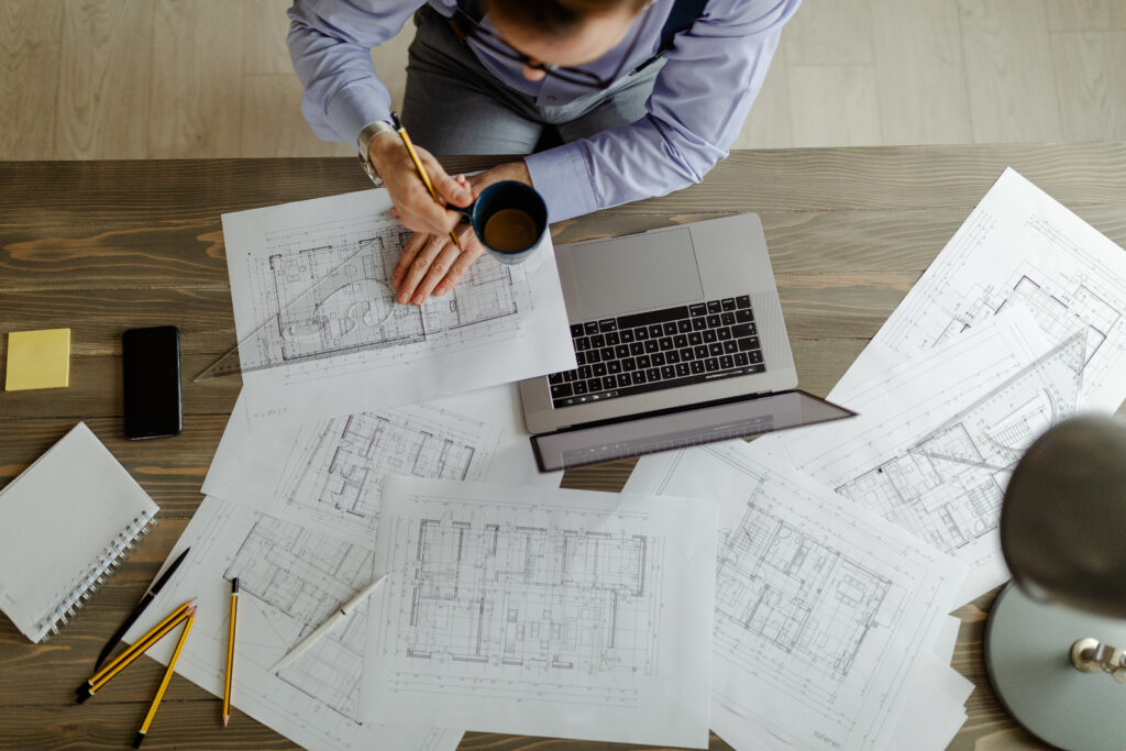 Overhead view of a male architect at a wooden studio desk reviewing building blueprint layouts, holding a coffee cup, and working next to an open laptop.