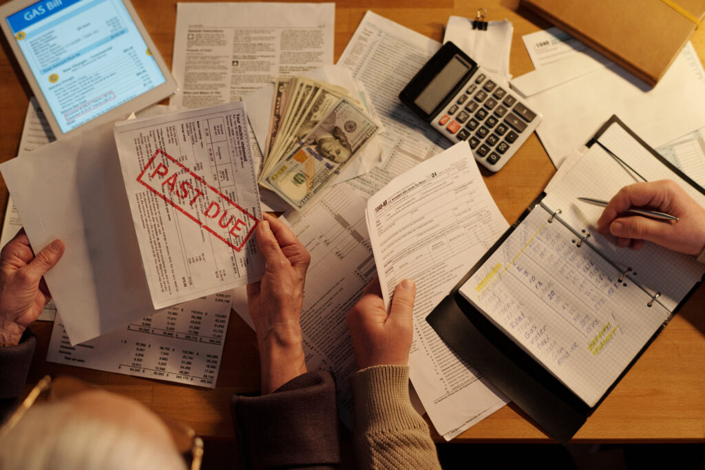 Top-down view of two people at a wooden desk reviewing financial documents, US cash, calculators, ledger books, and a bill stamped Past Due.