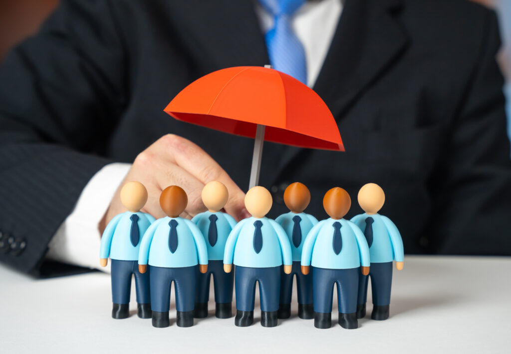 A wooden figure holding a protective red umbrella over a row of blue-uniformed employee figurines, representing business continuity and workforce protection