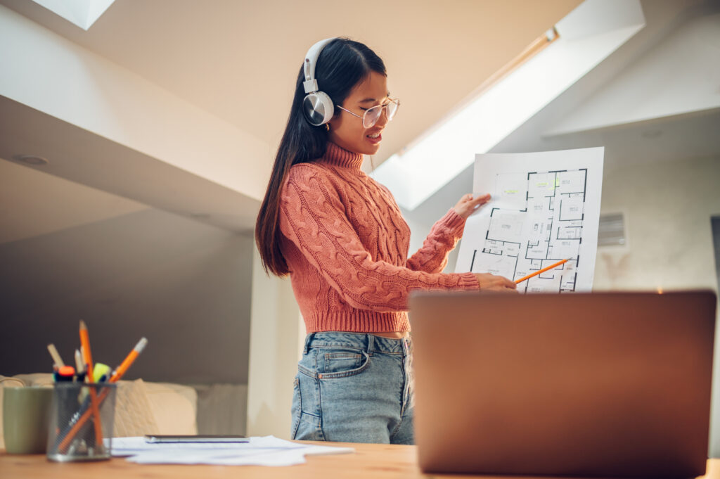 A remote female architect wearing headphones points at a printed floor plan during a video call on her laptop.