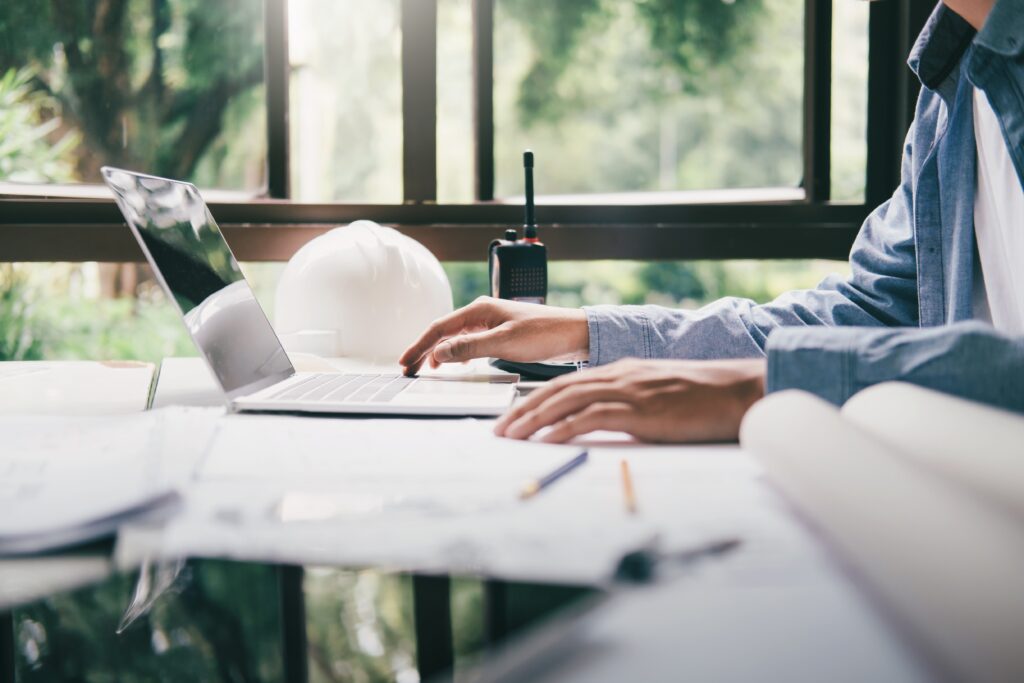 An AEC professional using a laptop at a desk with construction plans, blueprints, and a white hardhat