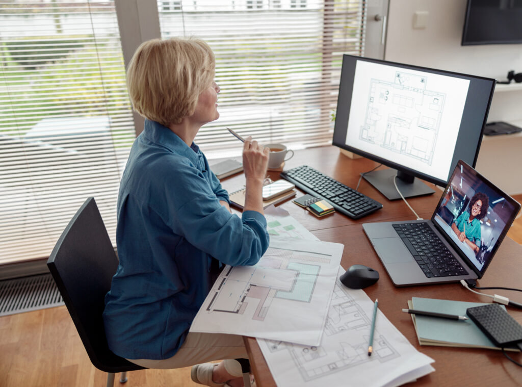 An architect works at a home office desk with a digital floor plan on a large monitor and a team video call on her laptop.