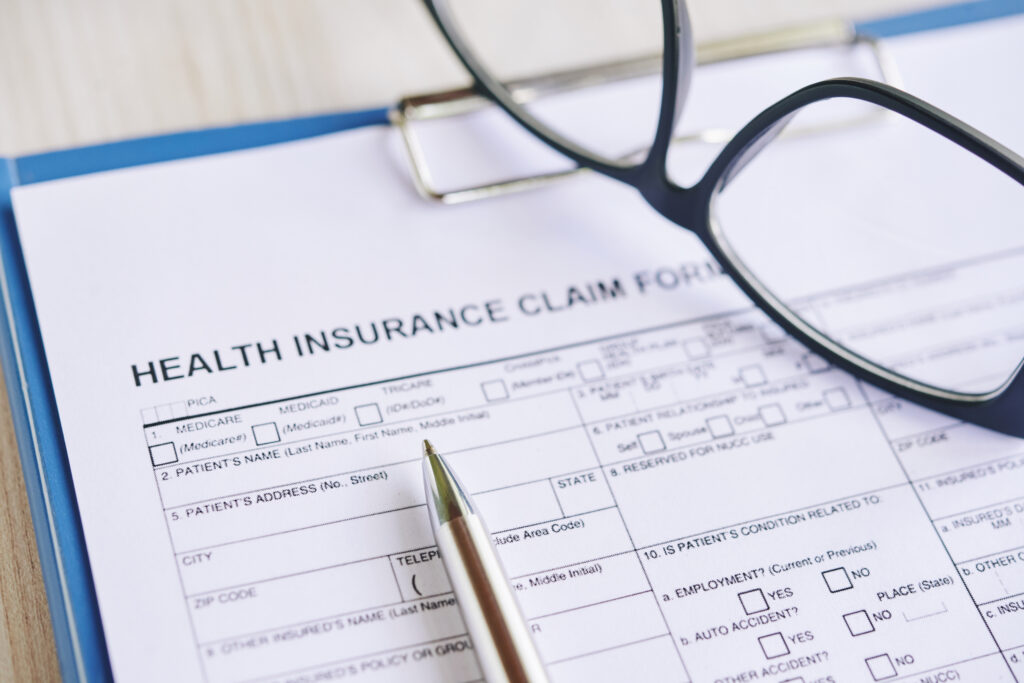 A printed health insurance claim form on a blue clipboard with a silver pen and reading glasses resting on top