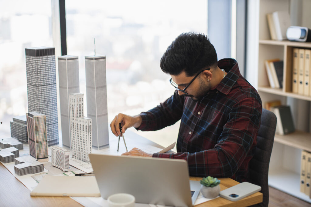 A male architectural engineer wearing a plaid shirt and glasses using a compass tool to measure scale models of city buildings and skyscrapers at an office desk next to a laptop.