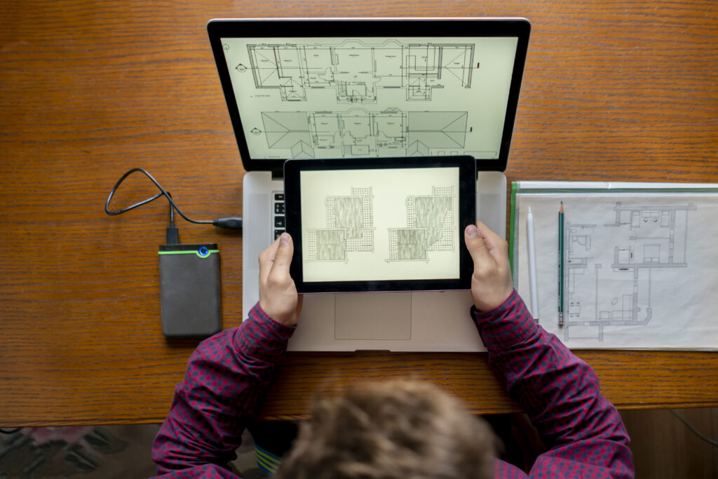 Overhead view of an architect comparing digital blueprints on a tablet and laptop next to printed schematics.