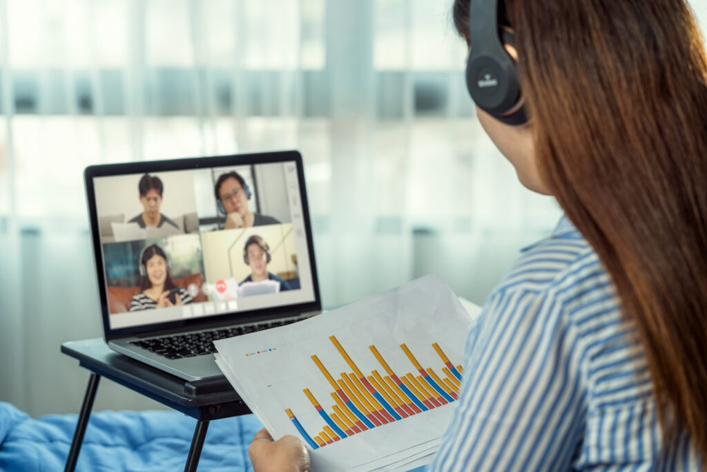 A remote architecture firm professional wearing headphones and reviewing a printed color bar graph during a multi-person video call on her laptop.