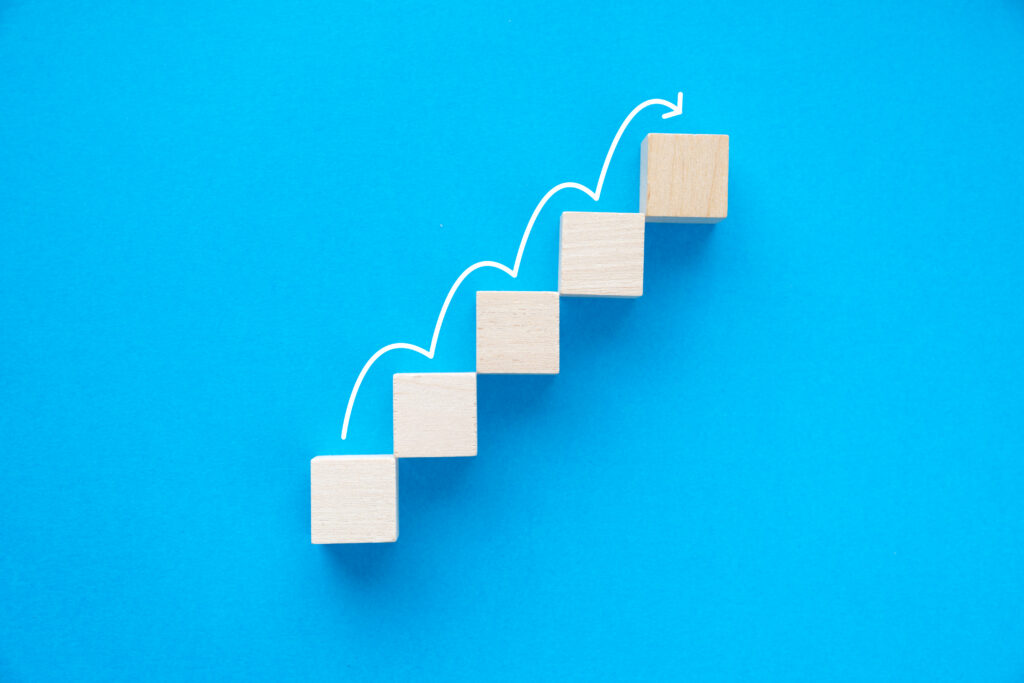 Wooden blocks arranged like a rising staircase on a vibrant blue background with an ascending curved white arrow above them.