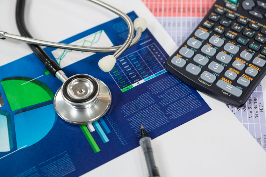 A medical stethoscope, calculator, and pen placed over printed financial charts and revenue analysis documents