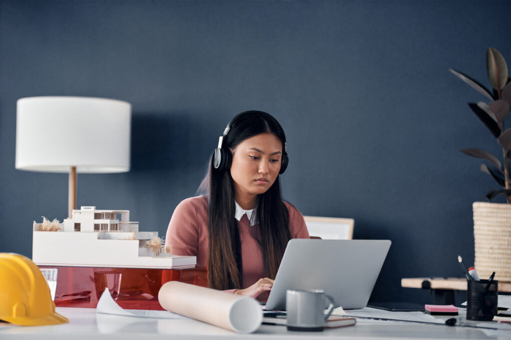 A female architectural professional wearing headphones and focusing closely on a laptop screen next to a 3D building model and a yellow hard hat on her office desk.