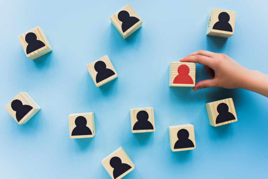 A hand selecting a red wooden block with a person icon among black blocks on a blue background, representing a dedicated credentialing specialist selected for specialized healthcare staffing.