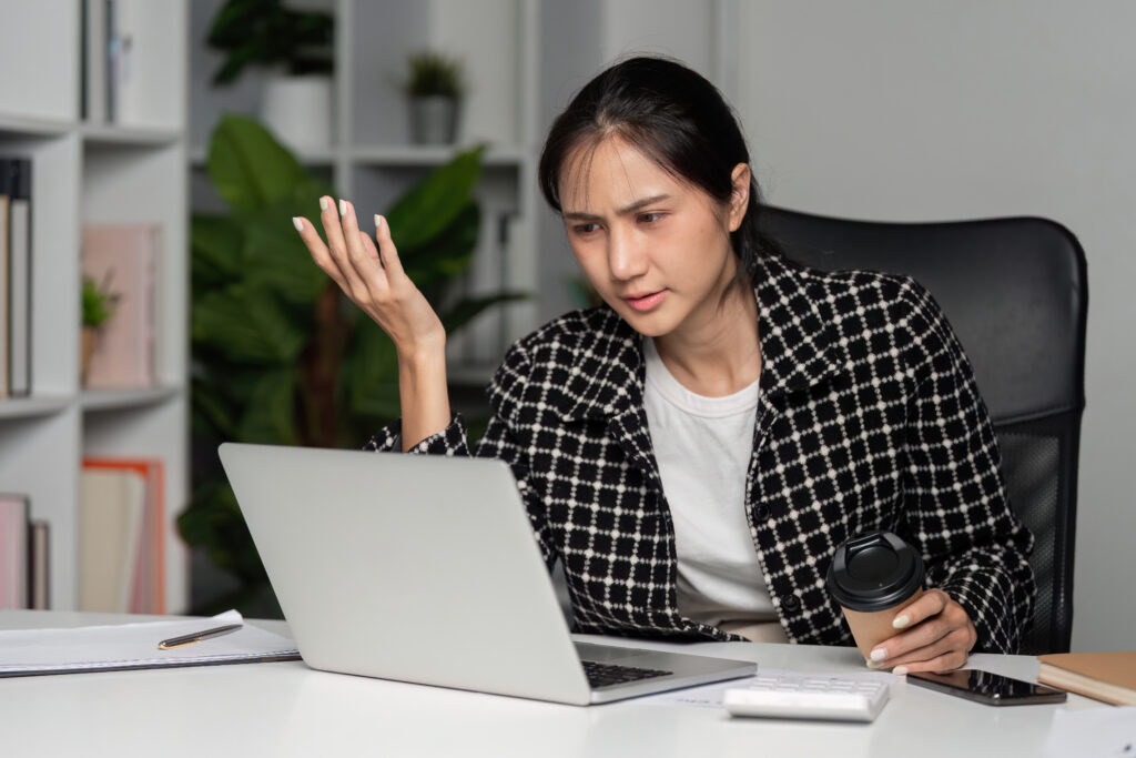 Stressed young female accountant sitting at a desk in front of a laptop holding a coffee cup, looking frustrated by financial work.