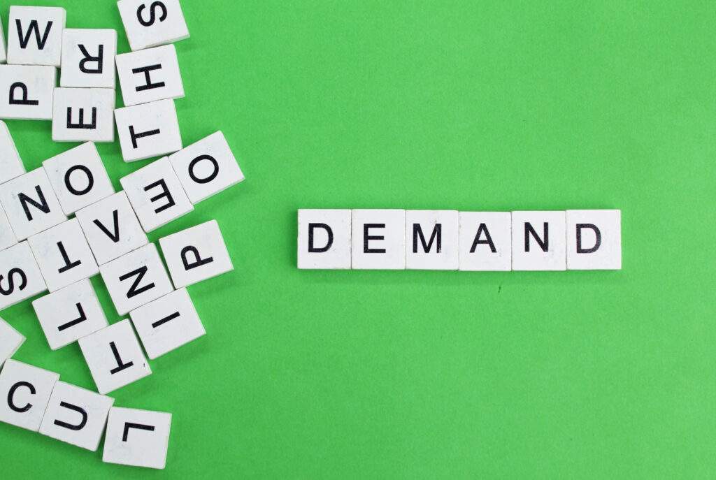 Wooden tile letters spelling out the word DEMAND on a vibrant green background surrounded by scattered letter tiles