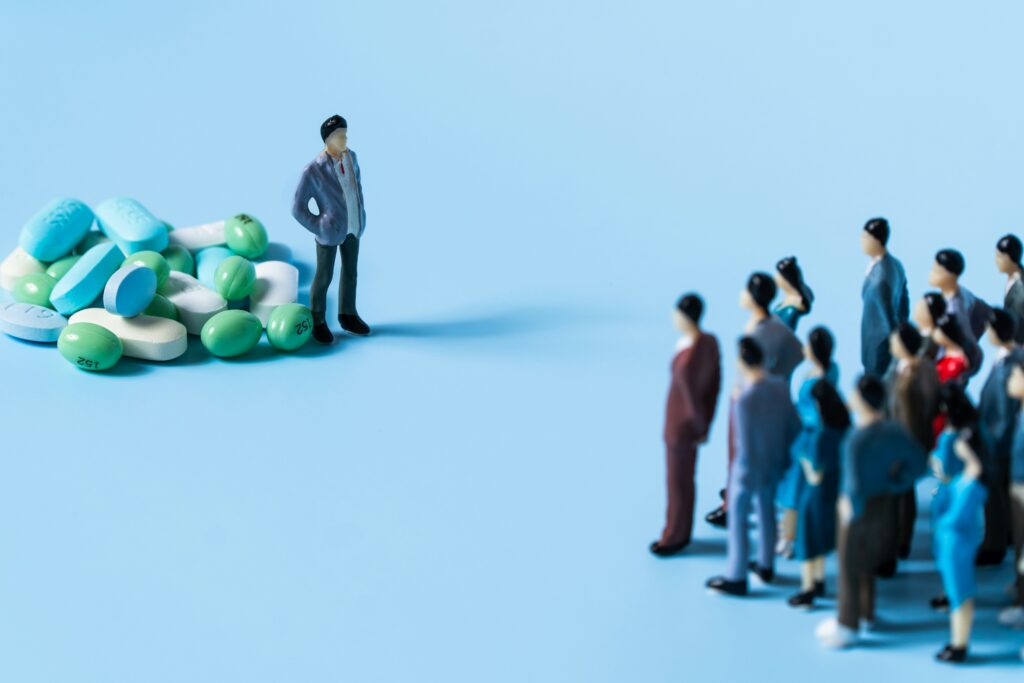 Miniature figure standing beside a pile of medication tablets, facing a crowd of small figures, illustrating payer policy changes affecting providers